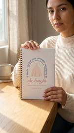 Woman holding a spiral-bound book titled 'she laughs' in a cozy indoor setting.
