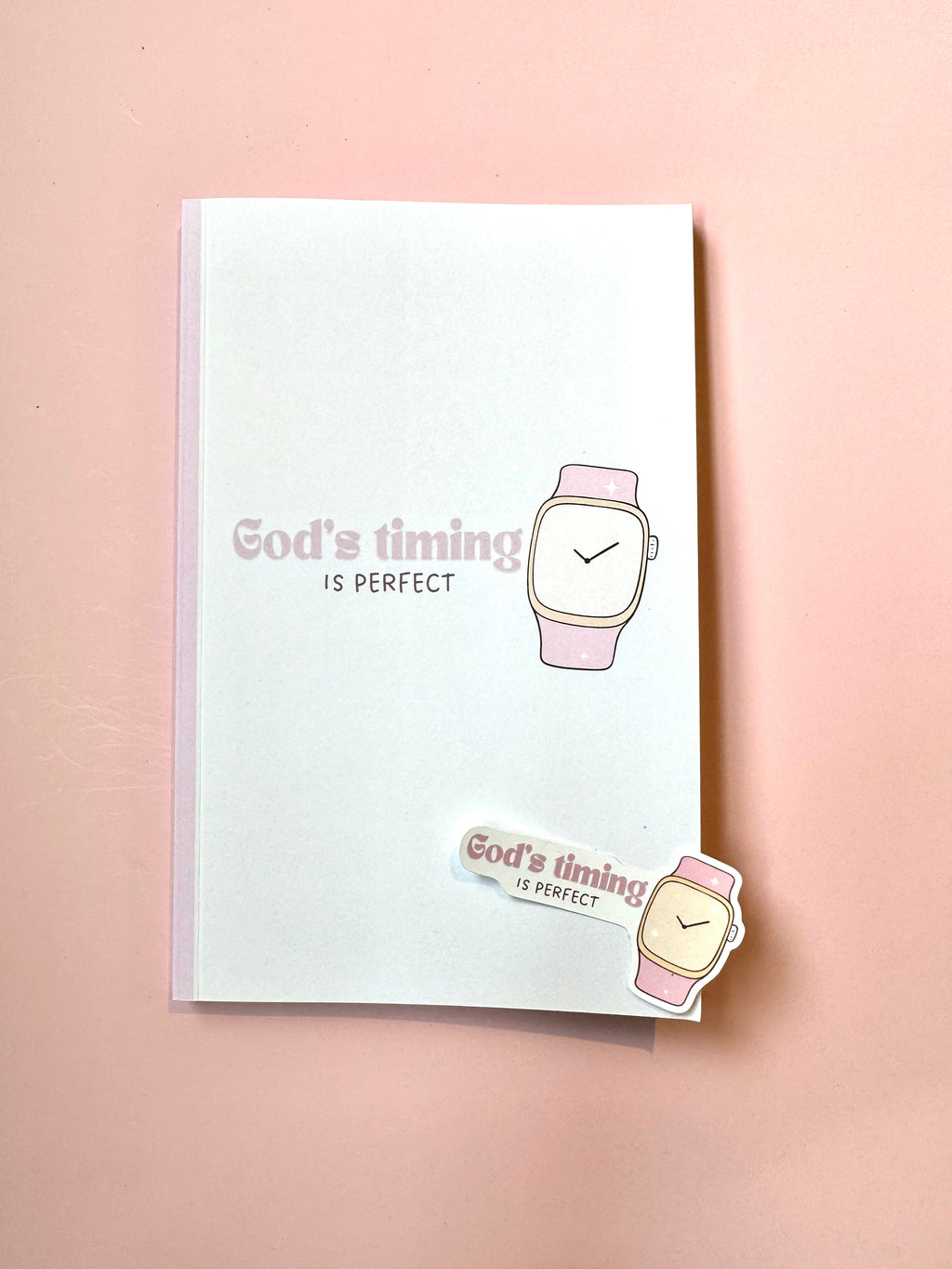 Notebook with 'God's timing is perfect' text and watch illustration on a pink background
